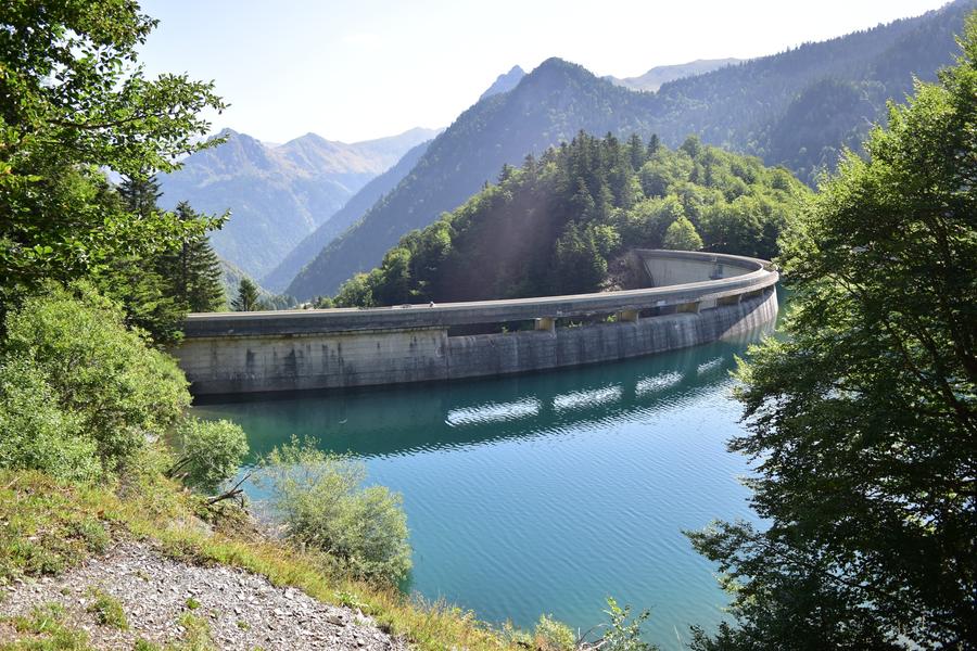 Photo d'un lac artificiel, l'eau est turquoise, on voit bien la structure du barrage en arc de cercle. Encore les montagnes en arrière-plan. 