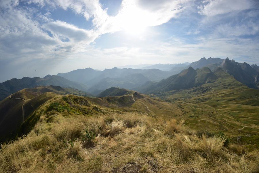 Photo panoramique (avec un objectf fish-eye) assez contrastée. On se situe sur un pic, un chemin serpente vers le bas, on voit bien les reliefs au loin. Le soleil tape déjà, malgré les nuages dans le ciel.
