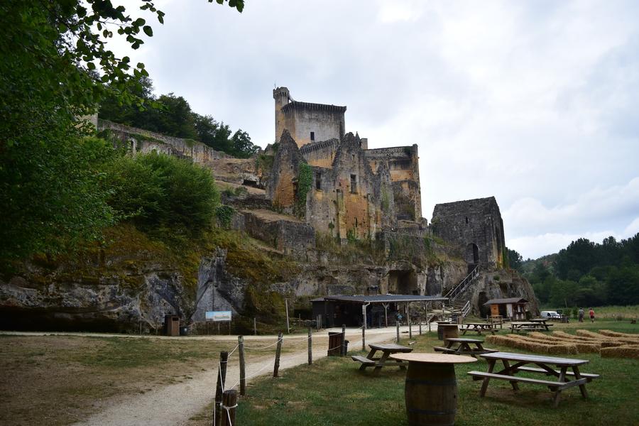 Photo du château de Commarque. On est en contrebas. Au premier plan, des tables de pique-nique. Plus loin, une falaise avecc des trous dedans. Sur la falaise, des bâtiments médiévaux, dont certains sont en ruines.
