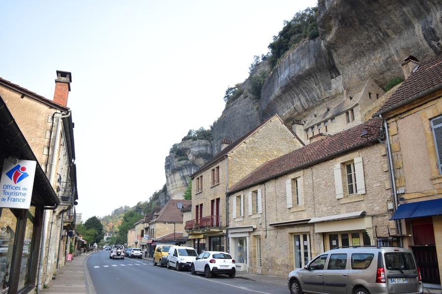Photo d'une rue de Tursac. À gauche, un panneau Office du tourisme ; au milieu la route ; à droite de belles maisons du coin avec leurs pierres beiges et leur toit en tuiles ; et derrière elles la falaise s'élève.